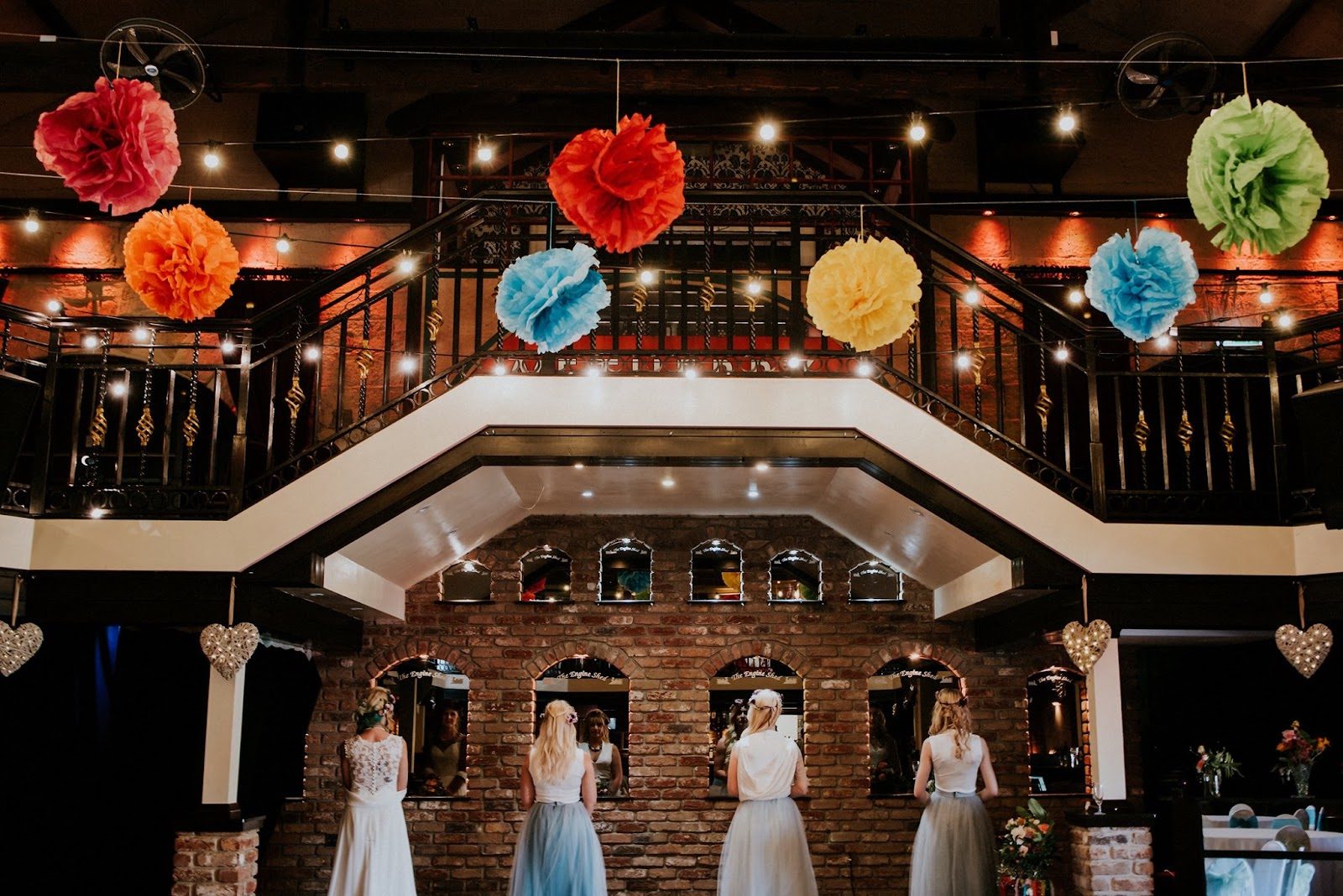 Bridesmaids at the bar beneath hanging paper pom-poms and the mezzanine