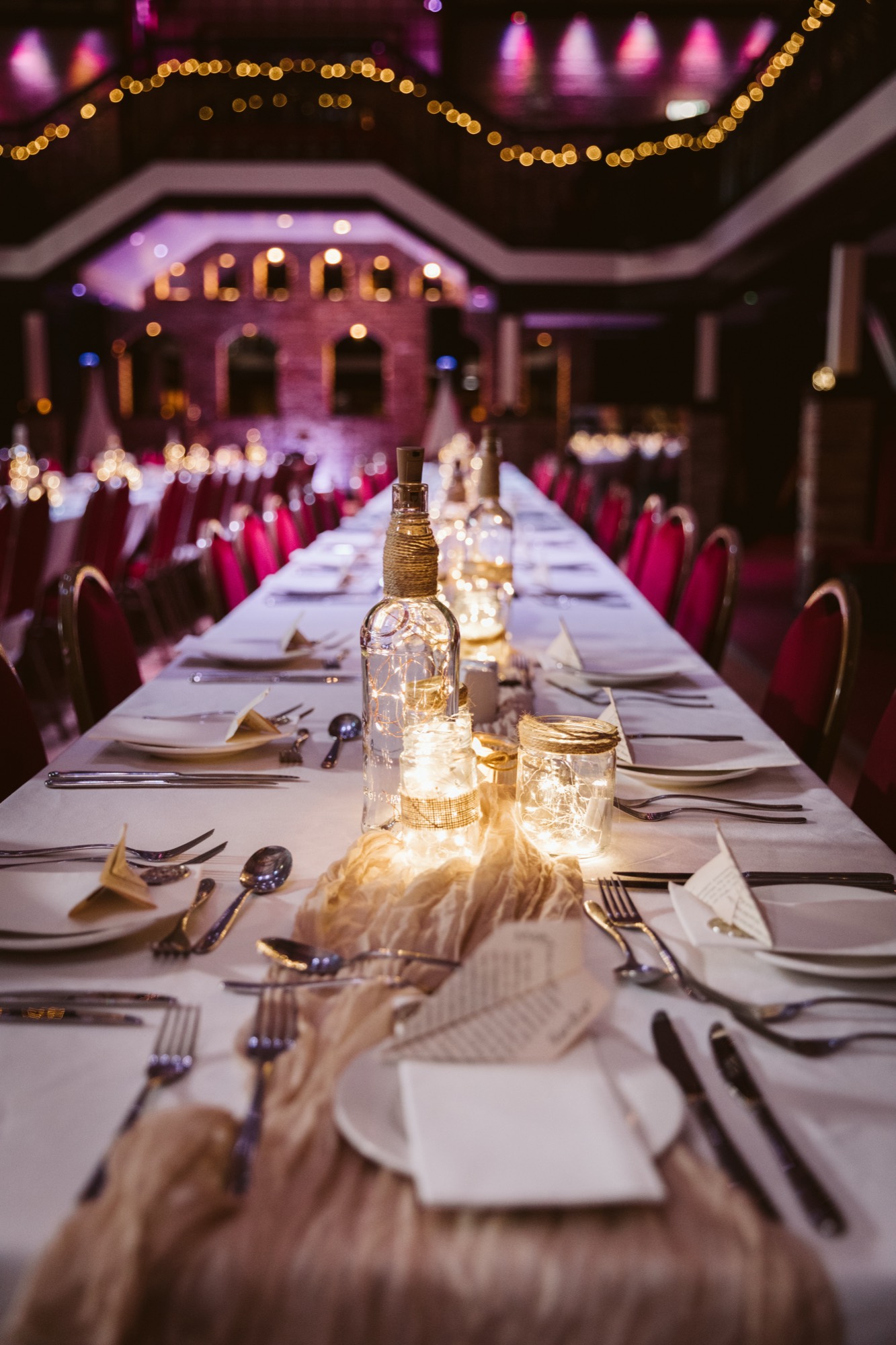 A long banquet table dressed with fairy-light jars, festoon lights above