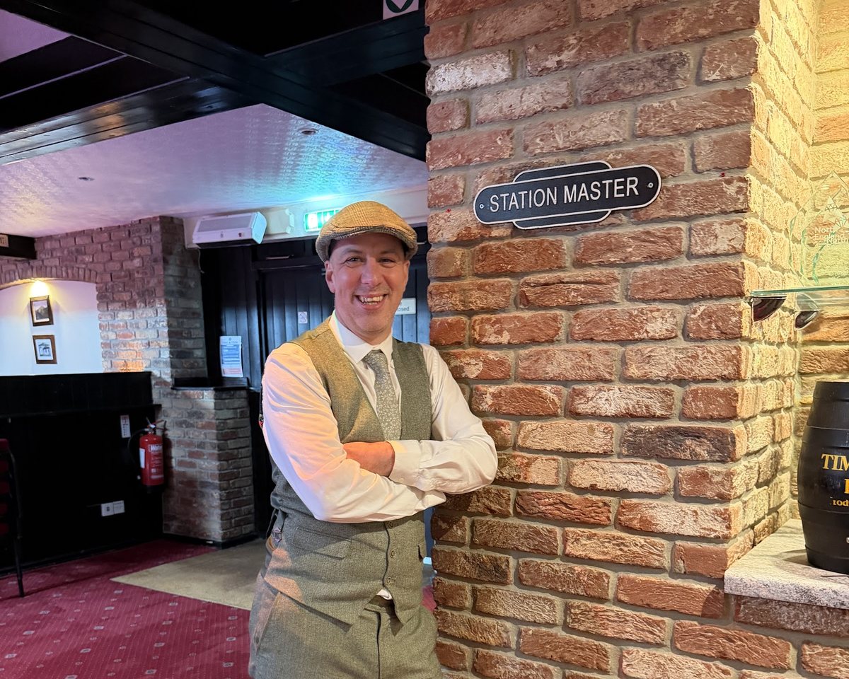 Lee, the wedding manager, in a flat cap and waistcoat beside the Station Master sign